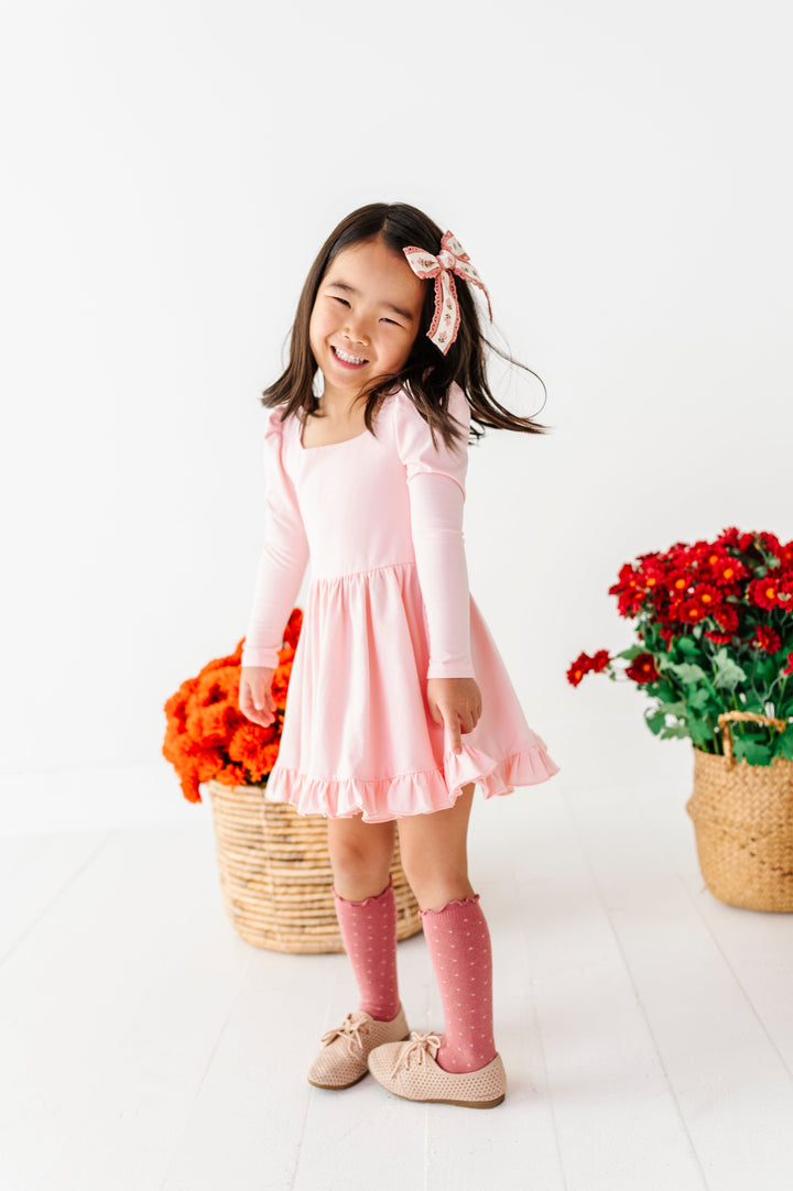 young girl twirling in front of baskets of fall flowers in pale pink long sleeve twirl dress, bright mauve dottie knee socks with matching floral hair bow