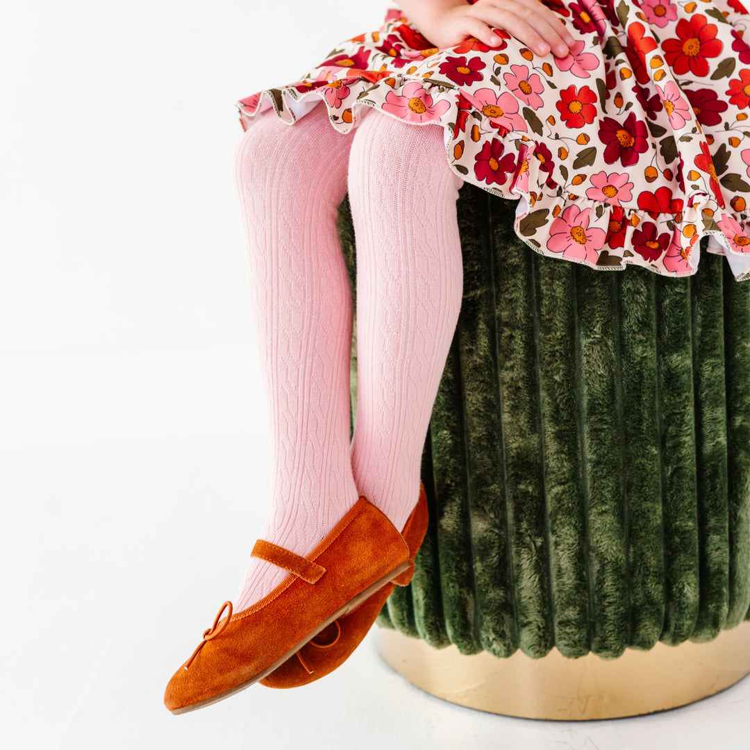 young girl sitting on green velvet stool wearing light pink cable knit tights paired with orange velvet flats and matching fall floral dress