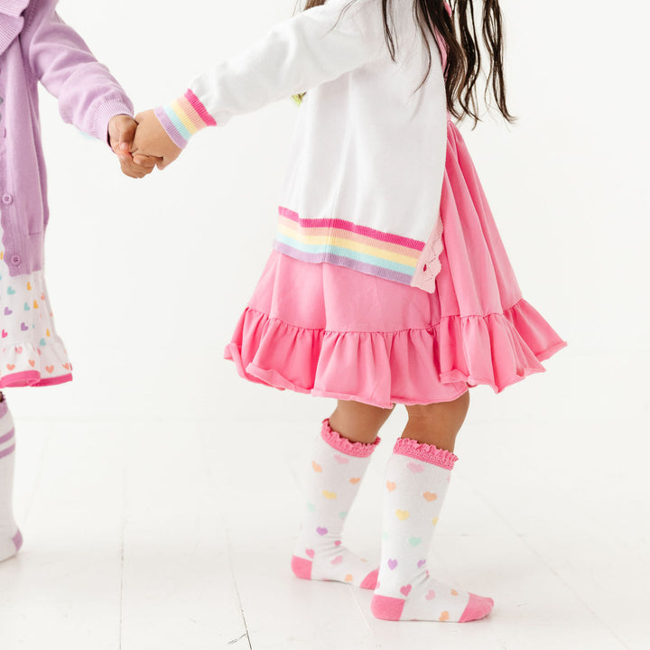 little girl in taffy pink dress wearing rainbow trim cardigan and rainbow heart knee high socks
