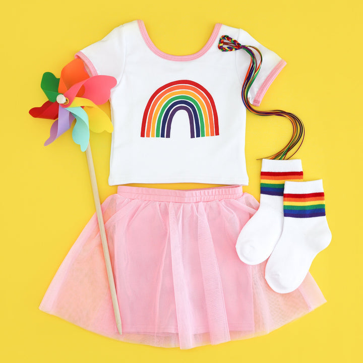 flat lay of fitted tee with rainbow design and pink binding paired with matching pink tutu shorts, rainbow midi socks, ribbon clip and pinwheel for pride day celebrations
