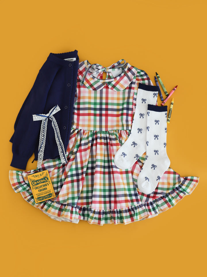 girl's colorful back to school flat lay outfit with collared rainbow gingham twirl dress and matching ribbon bow,  knee high socks with navy bow pattern and crayons