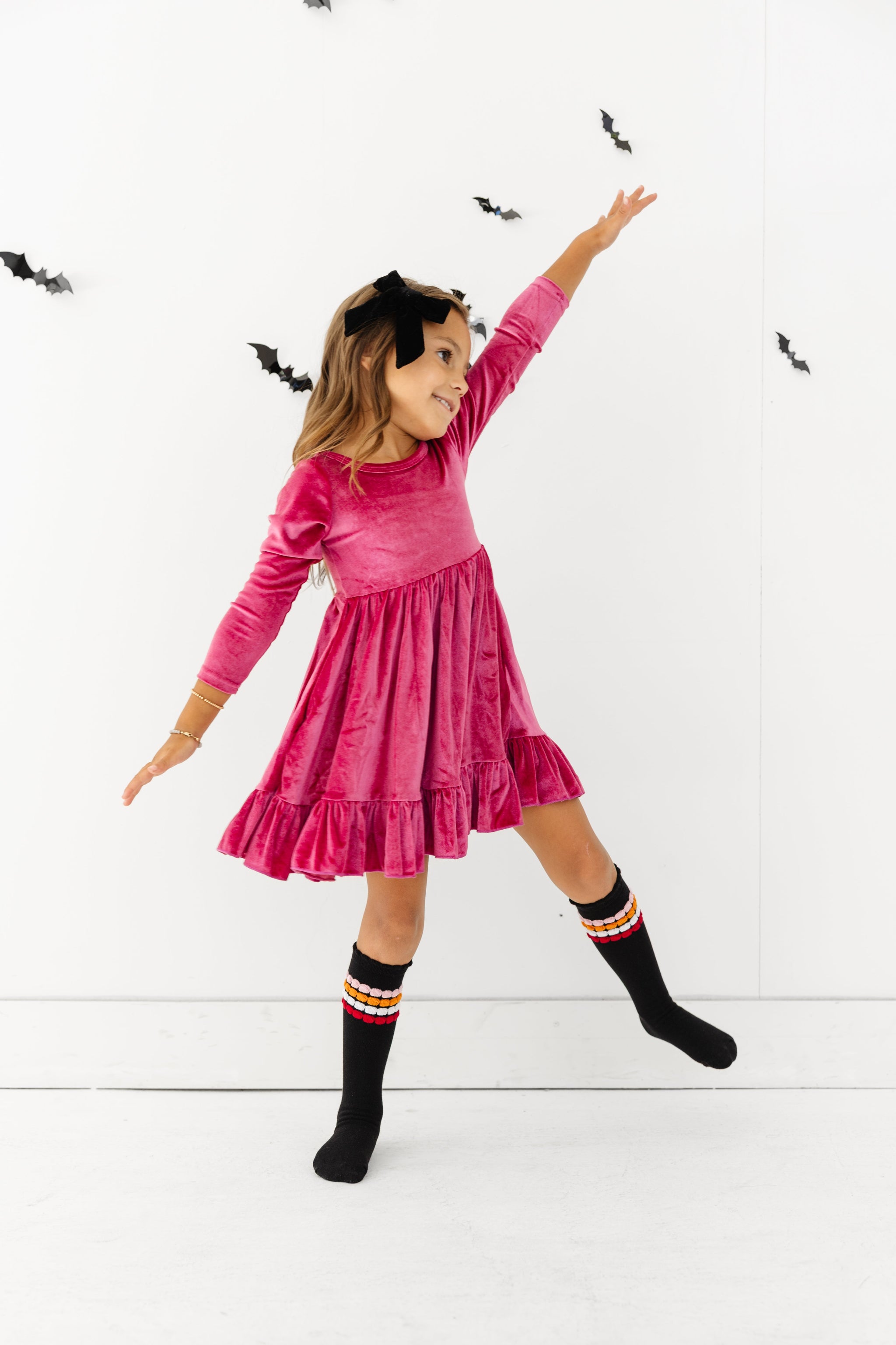 little girl in raspberry pink long sleeve velvet twirly dress with ruffle hem paired with black knee high socks with bubble stripe detail and black velvet bow