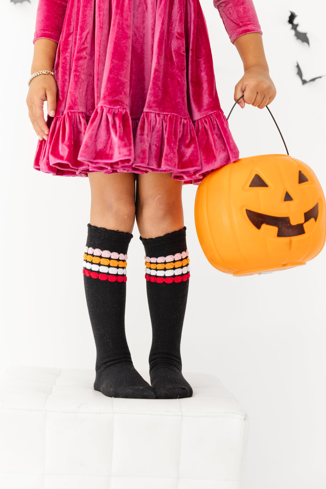 little girl holding pumpkin bucket in raspberry pink velvet charm dress and black knee highs socks with pink, orange and white bubble stripes