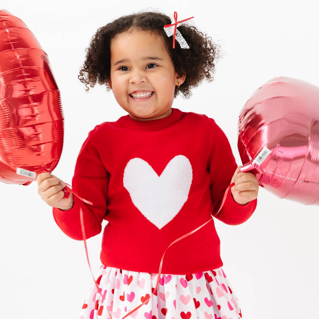 girl holding red and pink balloon wearing red sweater with white heart and matching valentine's day dress and bow