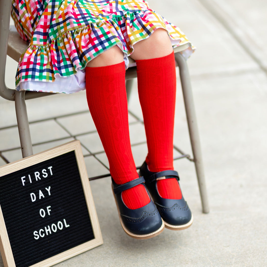 little girl sitting for first day of school photo wearing rainbow plaid dress and red cable knit knee high socks