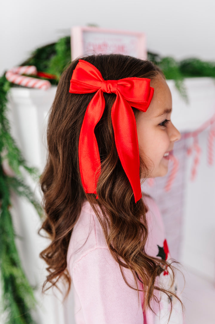 A young girl with long, wavy brown hair wearing a large, bright red satin bow headband, shown in profile against a festive holiday background with green garland and candy canes.