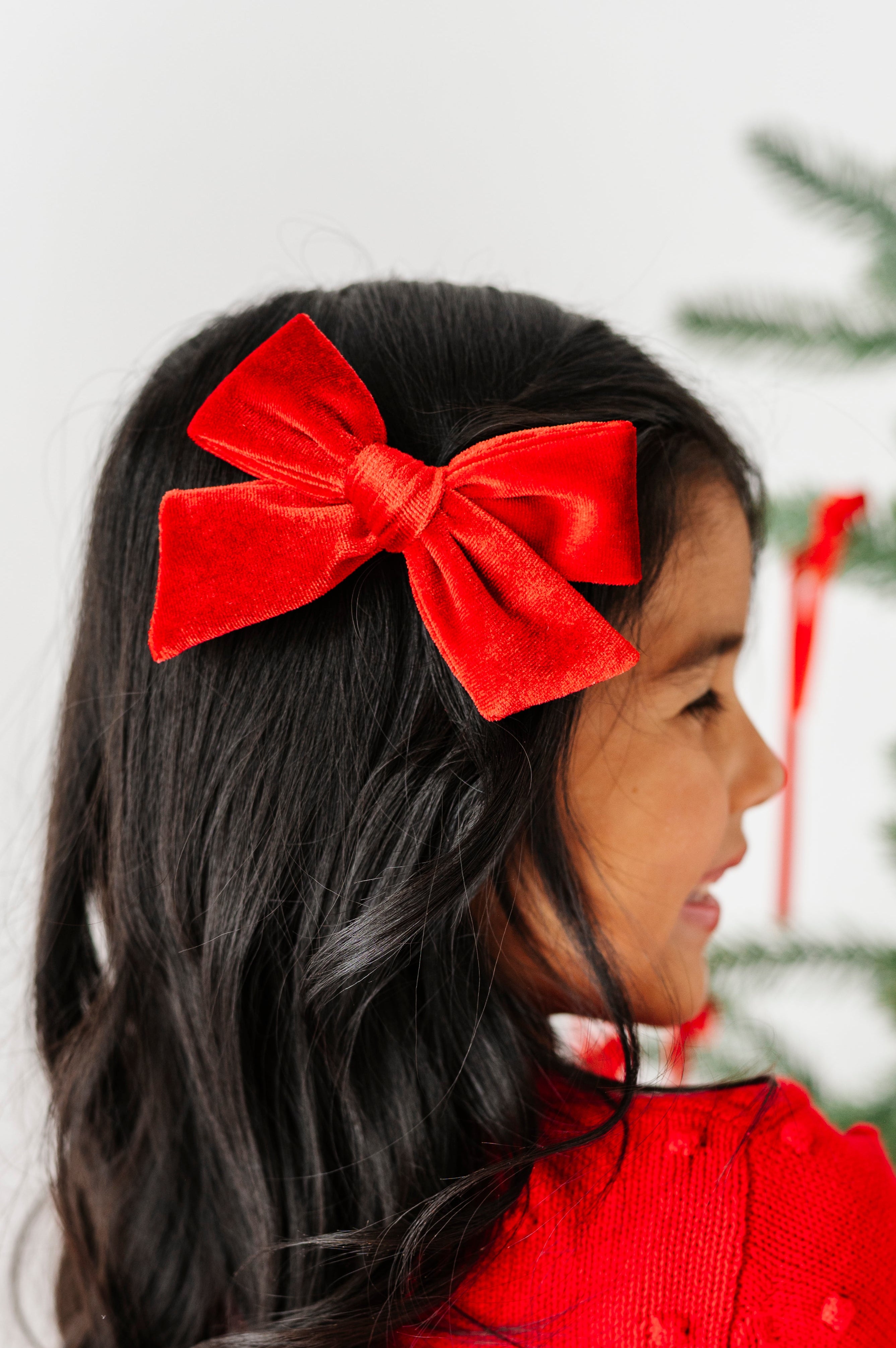 A young girl with long dark hair wearing a large red velvet bow headband and a red textured sweater, seen from the side as she looks at a Christmas tree with red ornaments.