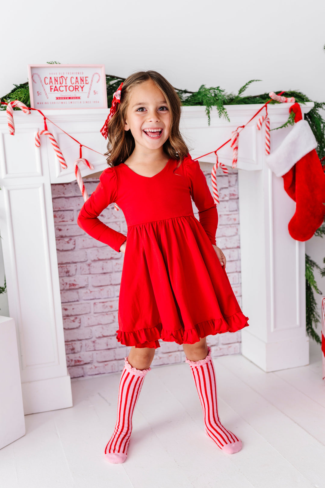A young girl stands in front of a white fireplace mantel decorated with green garland, candy canes, and a red Christmas stocking, wearing a bright red long-sleeve dress and red-and-white striped knee-high socks.