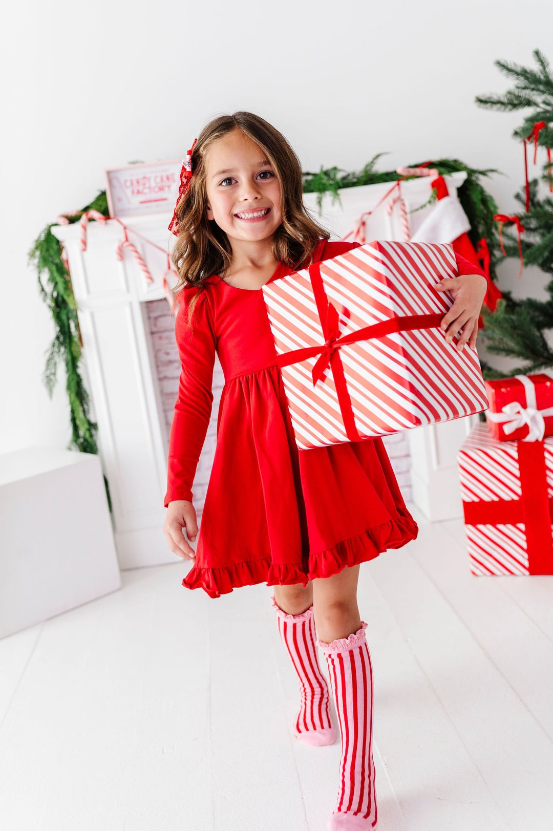 A young girl in a red long-sleeve dress and red-and-white striped knee-high socks holds a large red-and-white striped gift box with a red ribbon in a bright white holiday room with a Christmas tree and fireplace.
