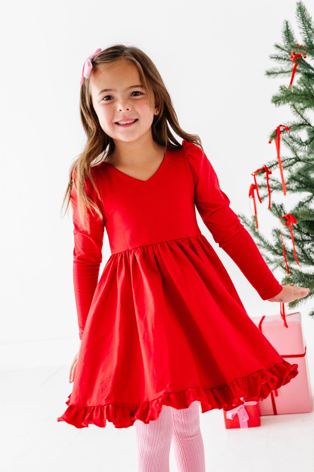 A young girl smiling and wearing a bright red long-sleeve dress with a ruffled hem, standing next to a decorated Christmas tree and holding a pink gift bag.