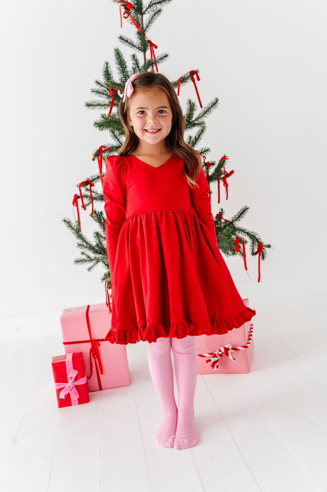 A young girl with brown hair stands in front of a decorated Christmas tree wearing a bright red long-sleeve dress, pink tights, and a pink bow headband. Pink gift boxes are wrapped around her legs.