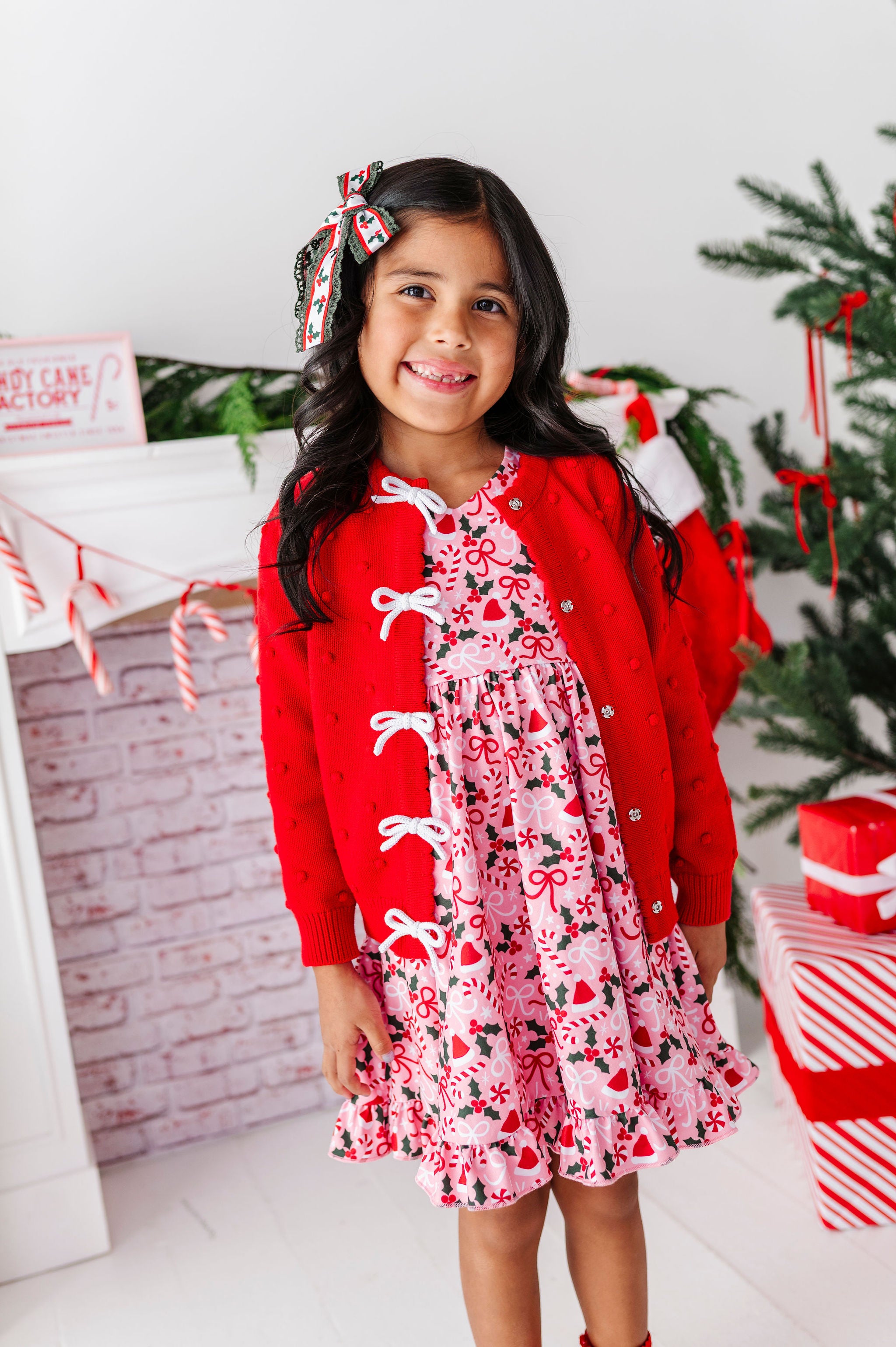 A young girl smiling in a festive holiday outfit, featuring a red knit cardigan with white bow details over a pink and red Christmas print dress, standing in front of a decorated Christmas tree and fireplace.