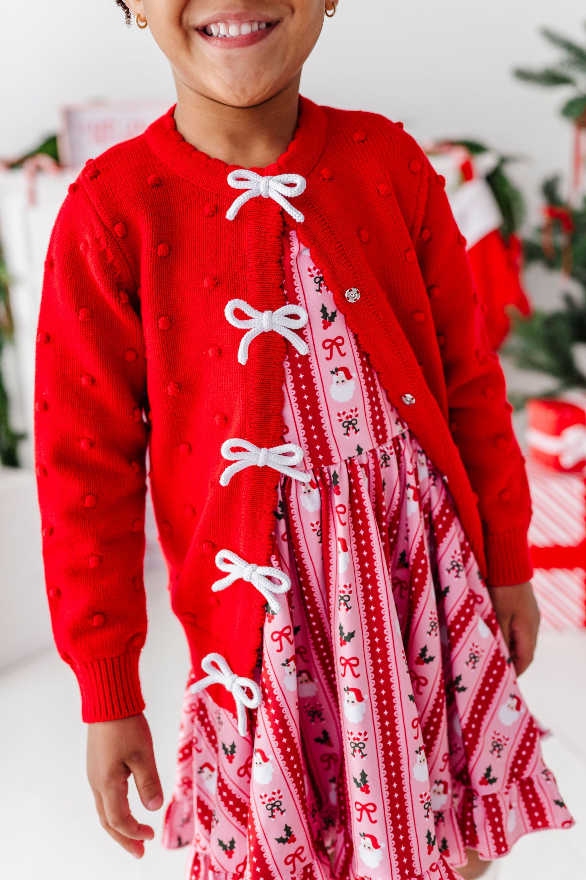 A child wearing a festive holiday outfit featuring a bright red cardigan with white bow details and a red and pink patterned dress underneath, set against a softly blurred Christmas-themed background.