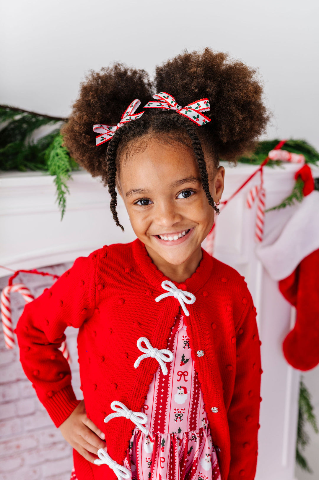 A young girl with two high-browed buns and braided sides wears a festive red knit cardigan with white lace-up front details over a holiday print top. She is smiling in front of a decorated holiday mantel with stockings, candy canes, and greenery.