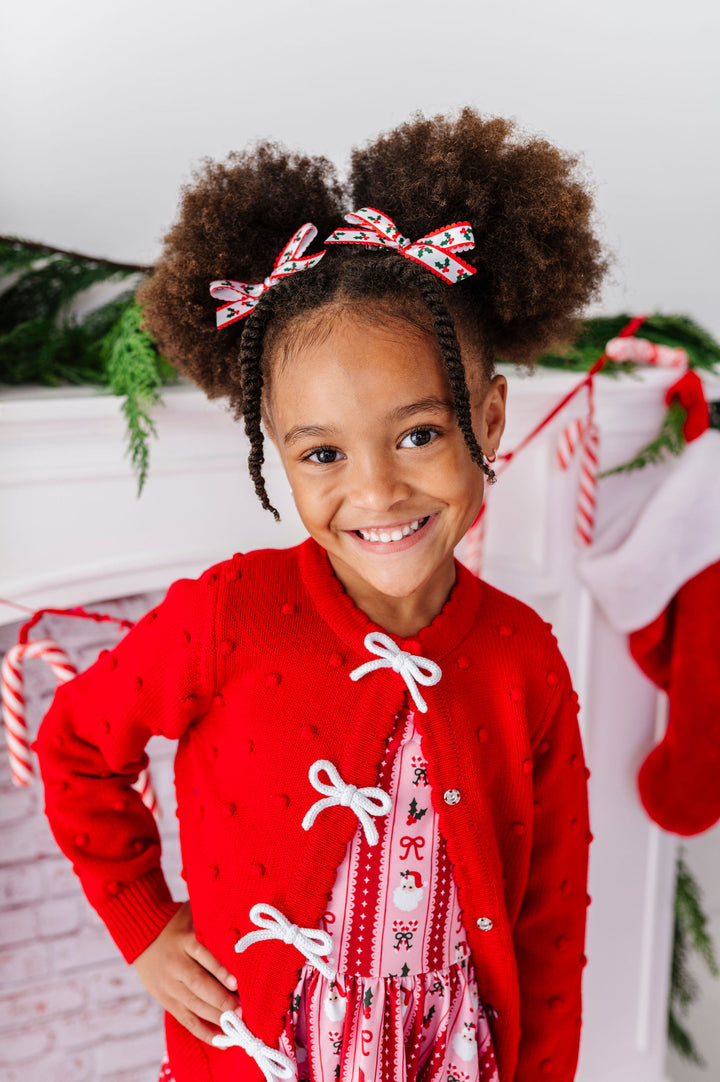 A young girl with two high-browed buns and braided sides wears a festive red knit cardigan with white lace-up front details over a holiday print top. She is smiling in front of a decorated holiday mantel with stockings, candy canes, and greenery.