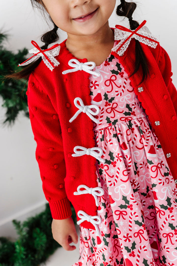 A young girl wearing a festive Christmas outfit, featuring a bright red cardigan with white bow details over a pink holiday print dress.