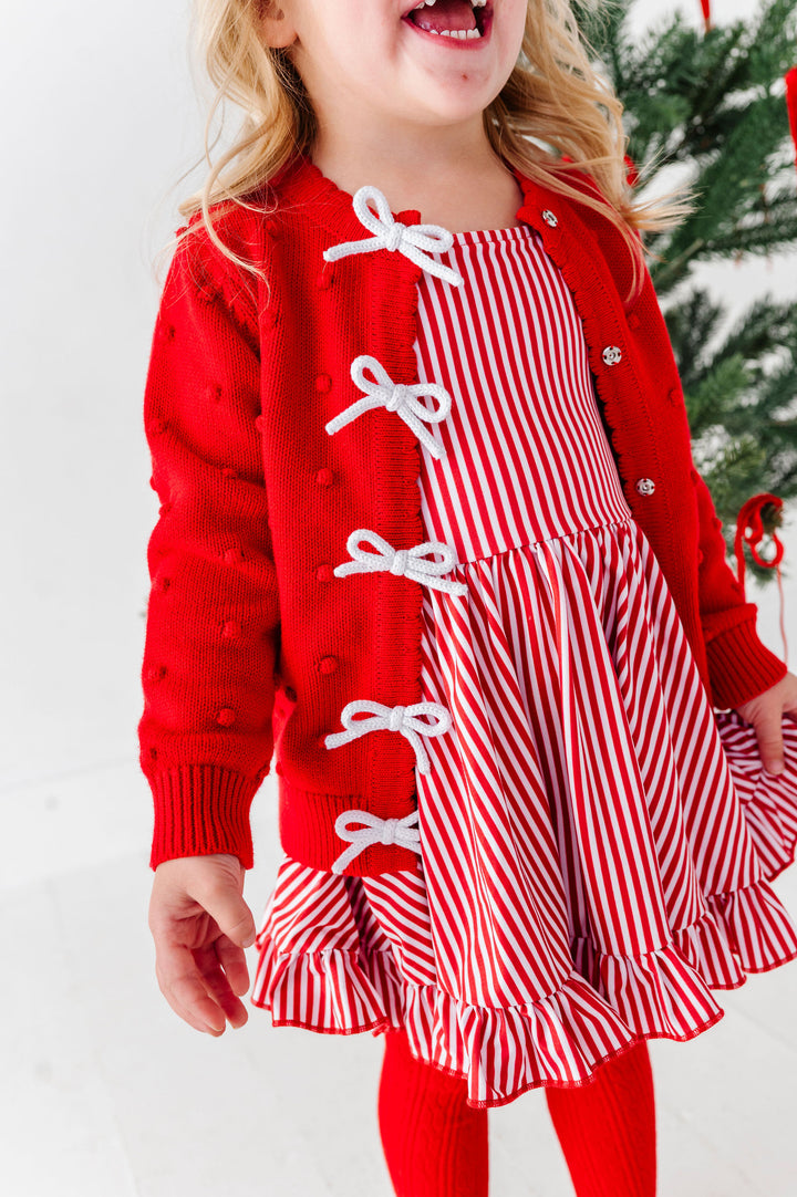 A young girl wearing a festive holiday outfit consisting of a red knit cardigan with white bow details and a red-and-white striped dress, standing in front of a Christmas tree.