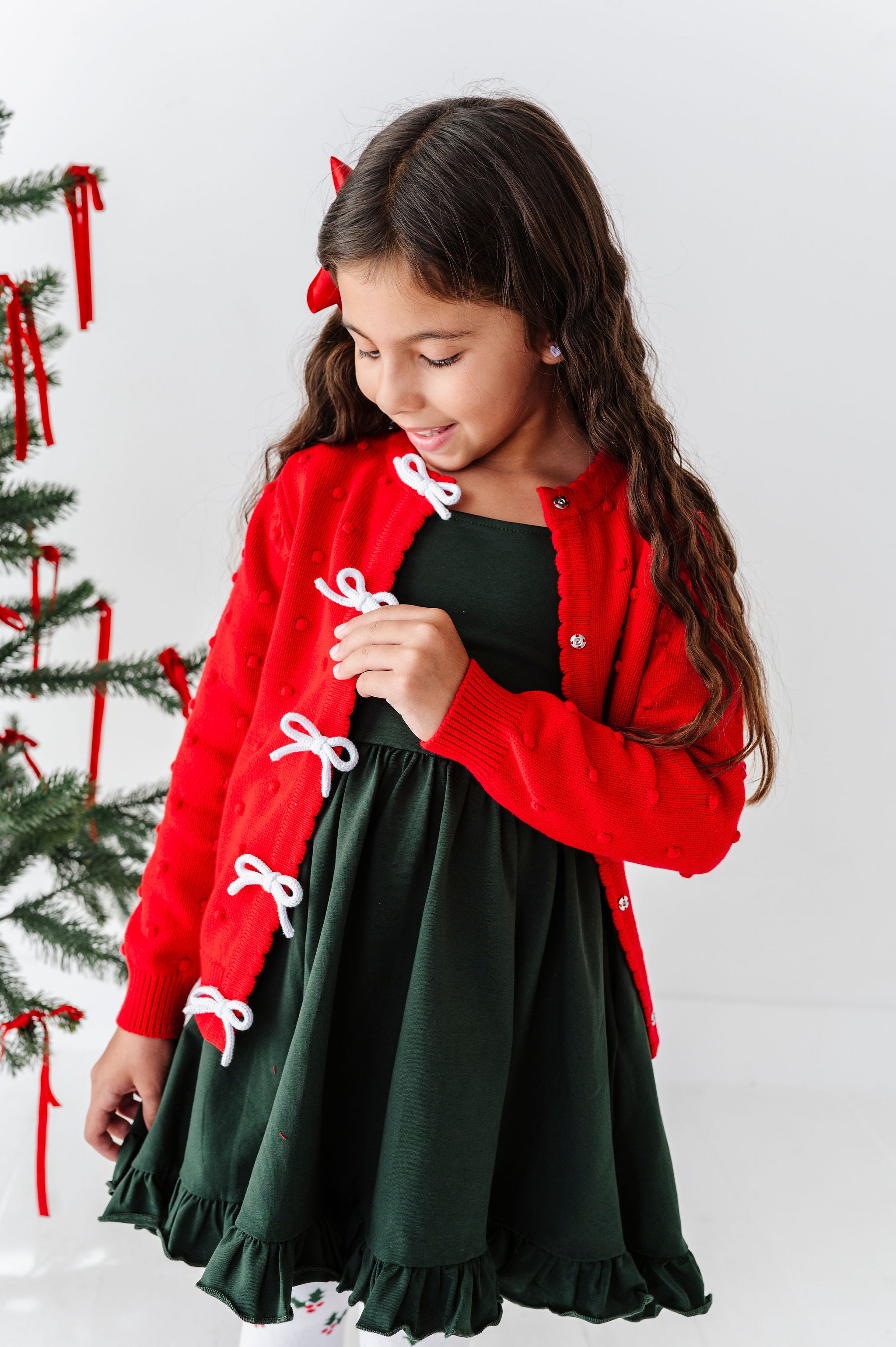 A young girl wearing a festive holiday outfit consisting of a red cardigan with white bow details and a dark green dress, standing next to a decorated Christmas tree against a white background.