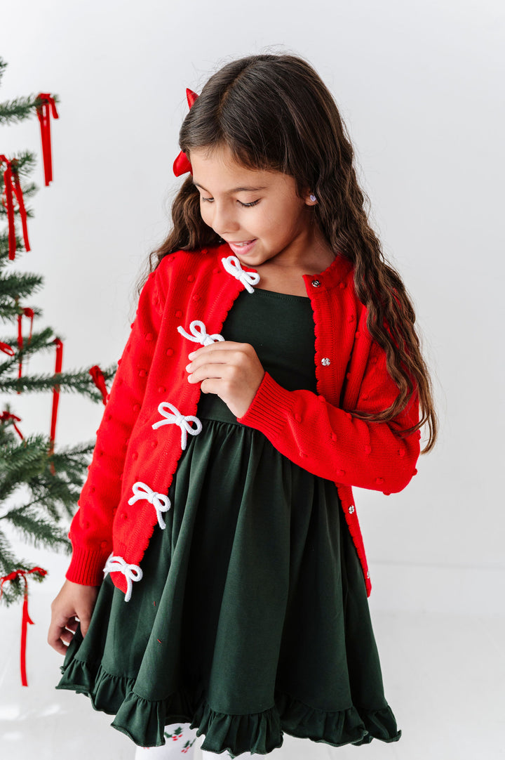 A young girl wearing a festive holiday outfit consisting of a red cardigan with white bow details and a dark green dress, standing next to a decorated Christmas tree against a white background.