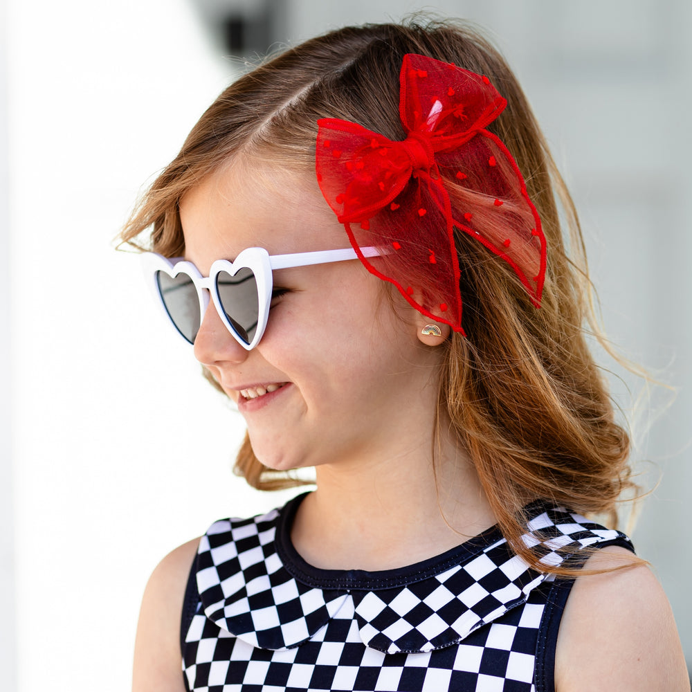 little girl wearing black and white checkered tank dress with white heart shaped sunglasses and a bright red sheer dot hair bow