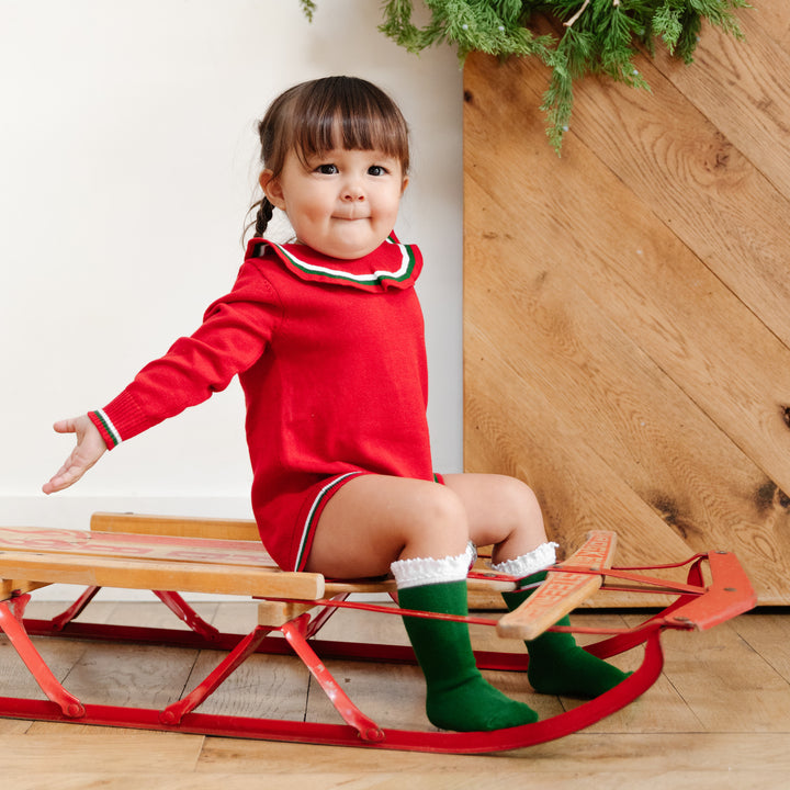 little girl sitting on sled in red christmas romper with adorable green and white knee high socks