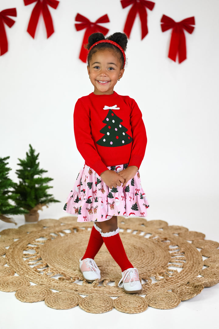 little girl in red sweater with Christmas tree design, matching pink Christmas print twirl dress and red knee highs with white lace trim 