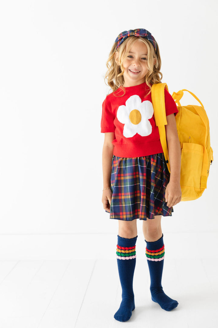 little girl holding yellow backpack wearing red cotton knit short sleeve sweater with flower design, navy plaid skort, matching plaid headband and navy knee highs with coordinating stripes