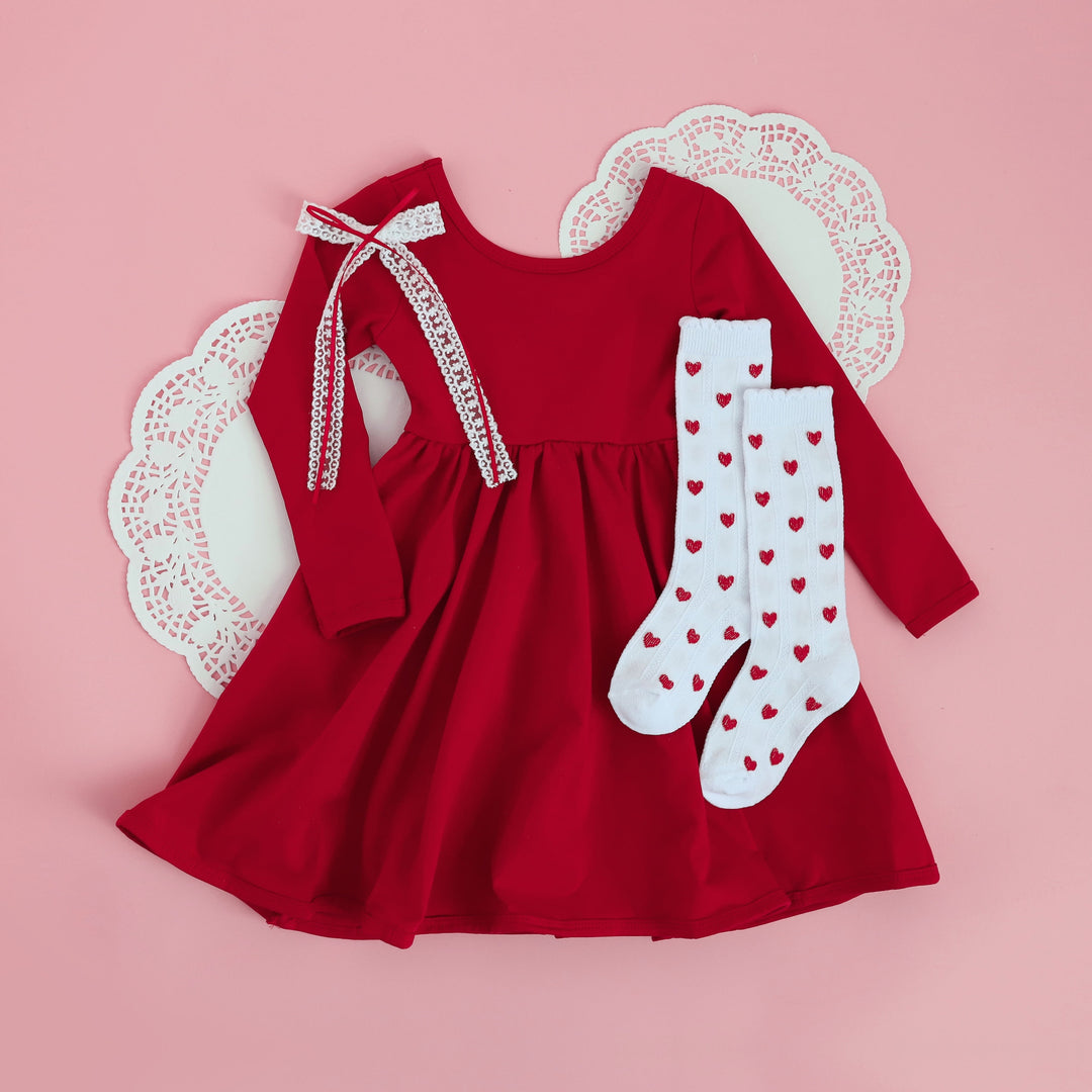 flat lay Valentine's day outfit for little girls featuring red stretch cotton long sleeve dress, red heart knee highs and white lace bow with red ribbon detail