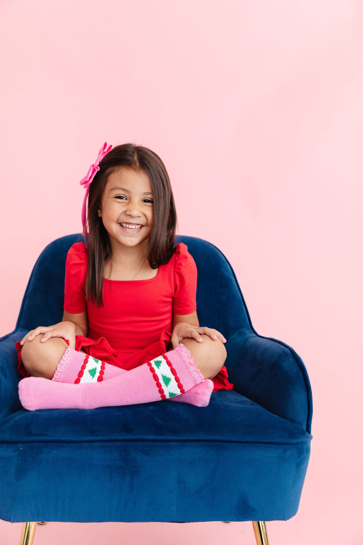 little girl sitting in classic red square neck twirl dress and matching pink knee highs with Christmas trees