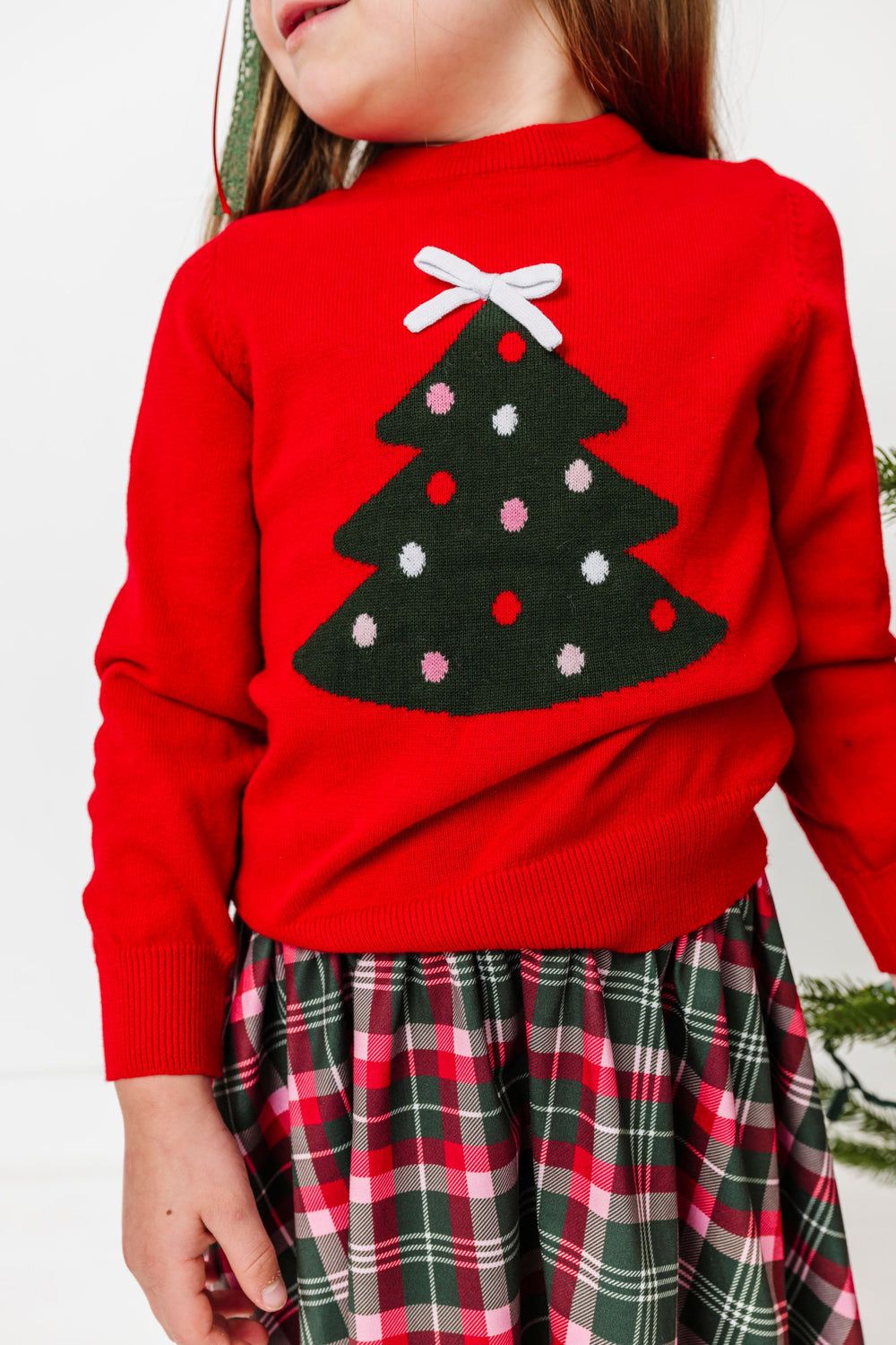 closeup of little girl in bright red knit sweater with forest green Christmas tree design decorated with red, pink and white dots and 3D white knit bow topper