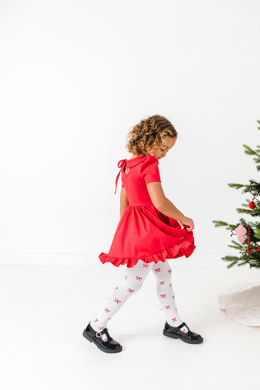 little girl in short sleeve red collared twirl dress paired with white tights with red bows by Christmas tree
