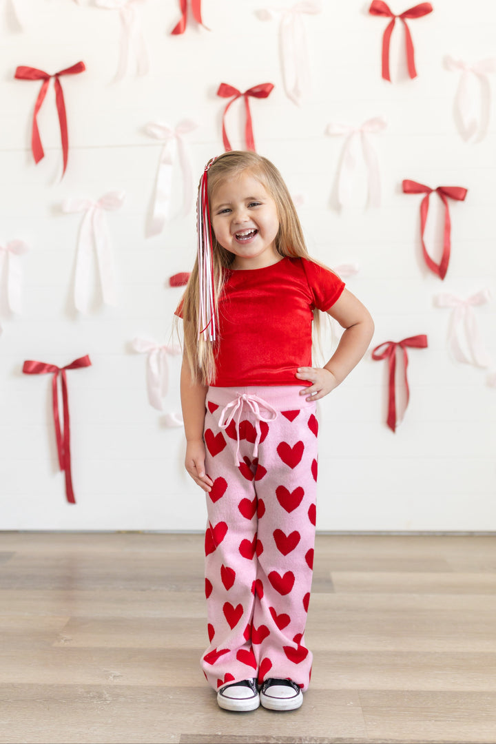 little girl in front of bow covered wall wearing bright red velvet fitted top paired with light pink cotton knit sweater pants with red heart design and matching ribbon hair clip