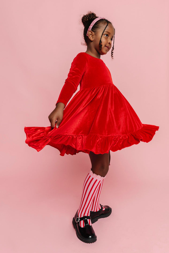 little girl twirling in bright red velvet long sleeve dress, pink sparkly headband and pink knee socks with vertical red stripe design for Christmas