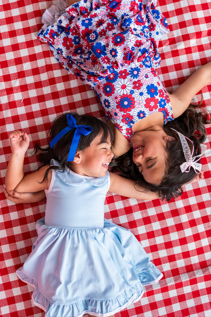 little girls on red gingham picnic blanket in red, white and blue floral twirl and light blue sleeveless twirl with matching hair bows