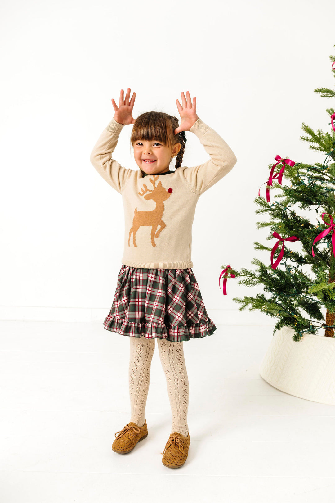 child by Christmas tree in cream pullover sweater with red nose reindeer design, matching green plaid Christmas dress and cream open knit tights