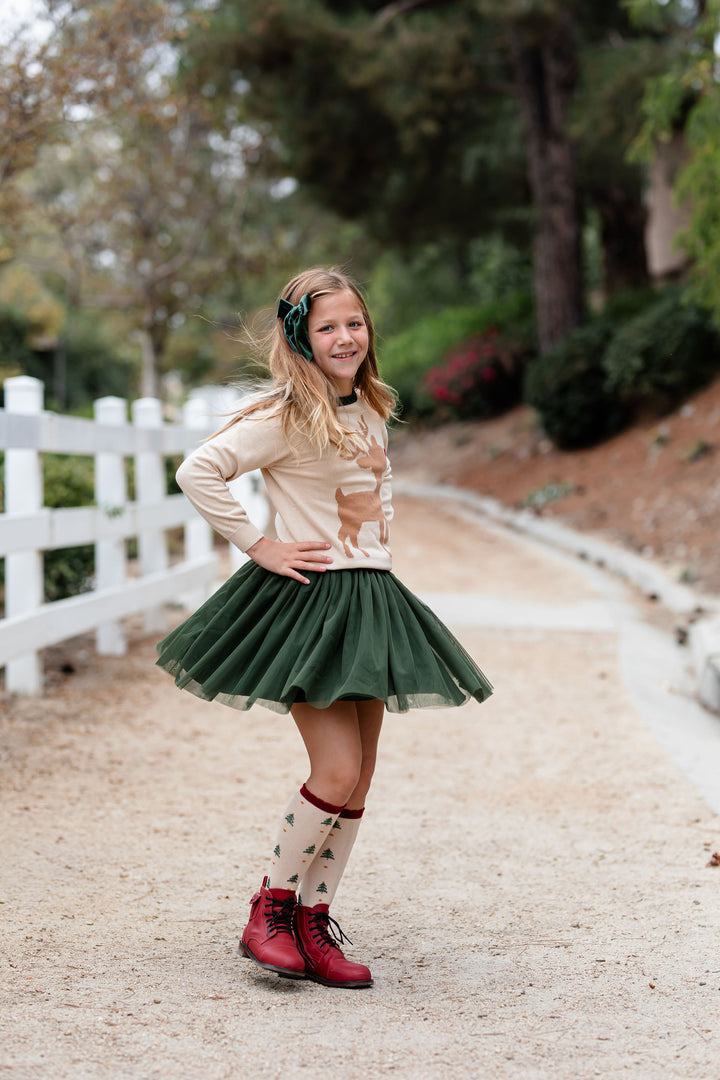 girl twirling on forest trail wearing cream reindeer print pullover knit sweater paired with forest green tulle skirt, matching velvet bow and Christmas tree print knee high socks with red boots