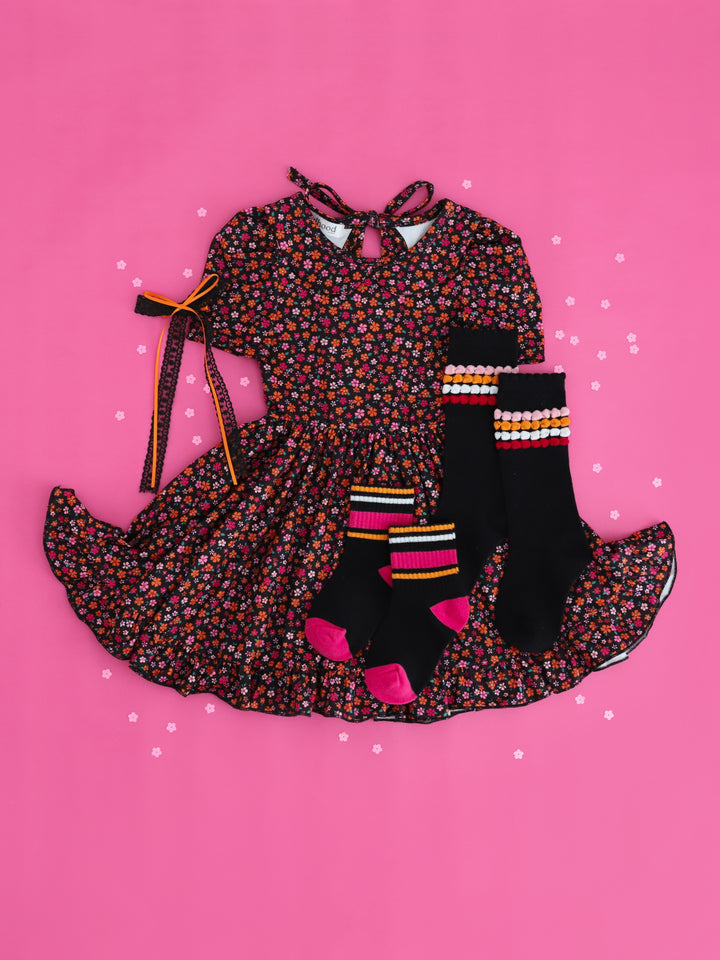 girls styled flat lay on hot pink with flower confetti featuring black collared twirl dress with petite orange and pink flower print paired with matching orange, black and pink socks and hair bow for Halloween vibes