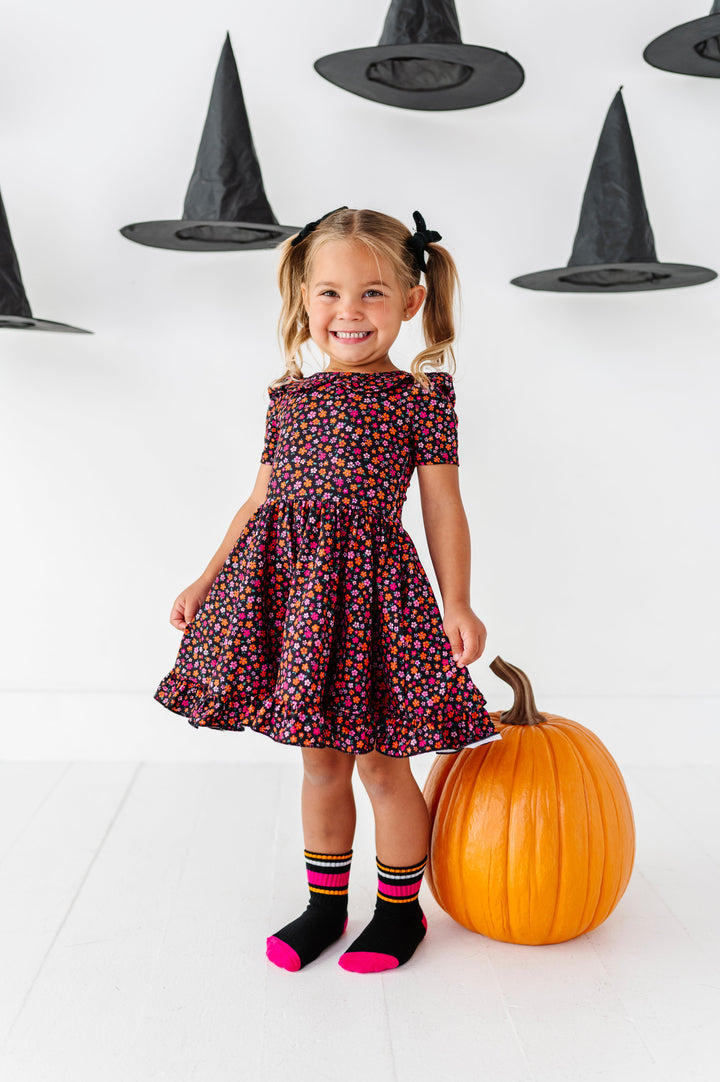 little girl standing by large pumpkin and surrounded by floating witch hats in black collared twirl dress with pink and orange petite flower print and matching striped crew socks