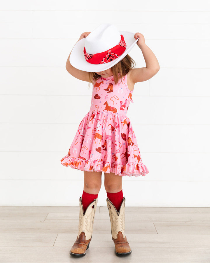 little girl in sleeveless pink rodeo print twirl dress wearing white cowgirl hat with red bandana, red knee highs and boots