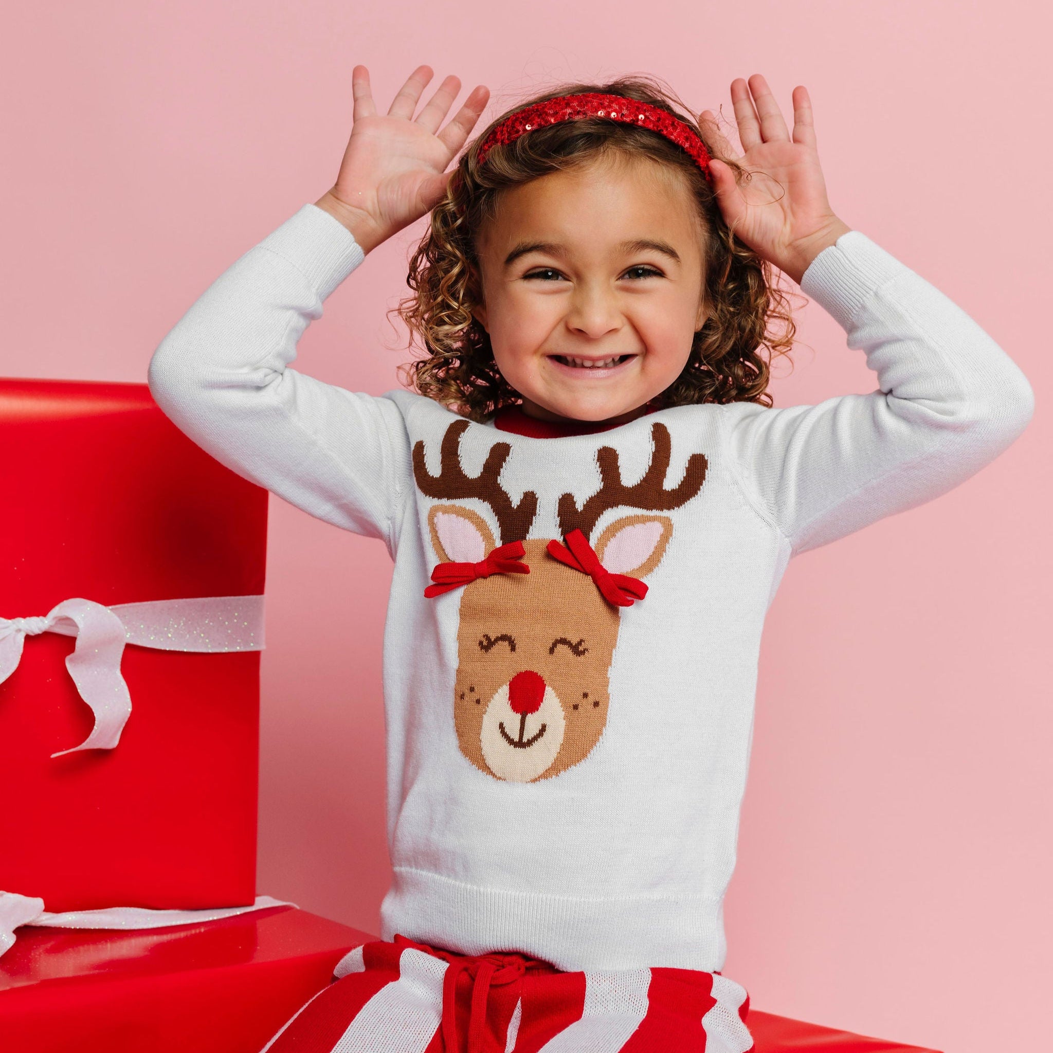 little girl sitting on Christmas gifts in white sweater with reindeer design, red sequin headband and pink and red striped sweater pants