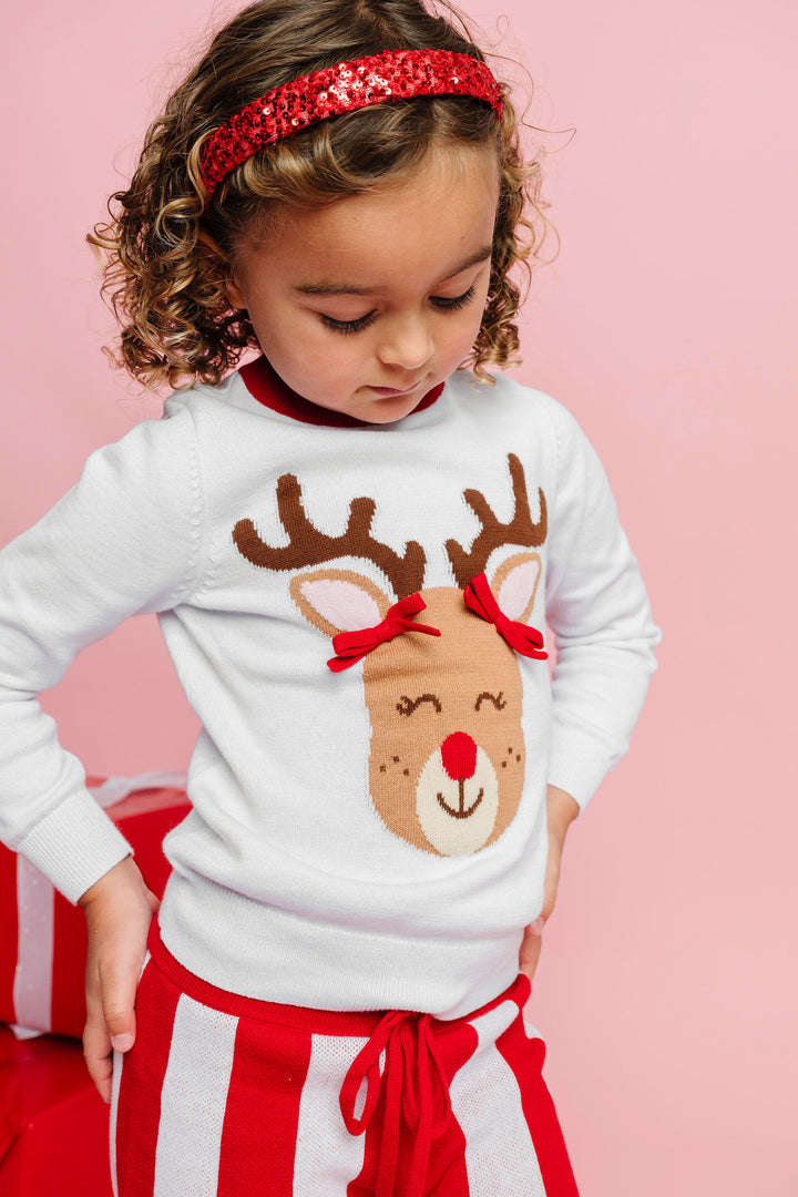 little girl in white knit sweater with reindeer wearing red pigtail bows paired with sparkly red headband and striped sweater pants