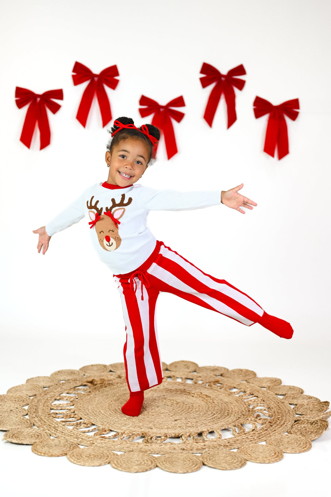 little girl in white pullover sweater with Rudolph design wearing 3D red pigtail bows paired with red and white striped sweater pants and red yarn pigtail bows for Christmas