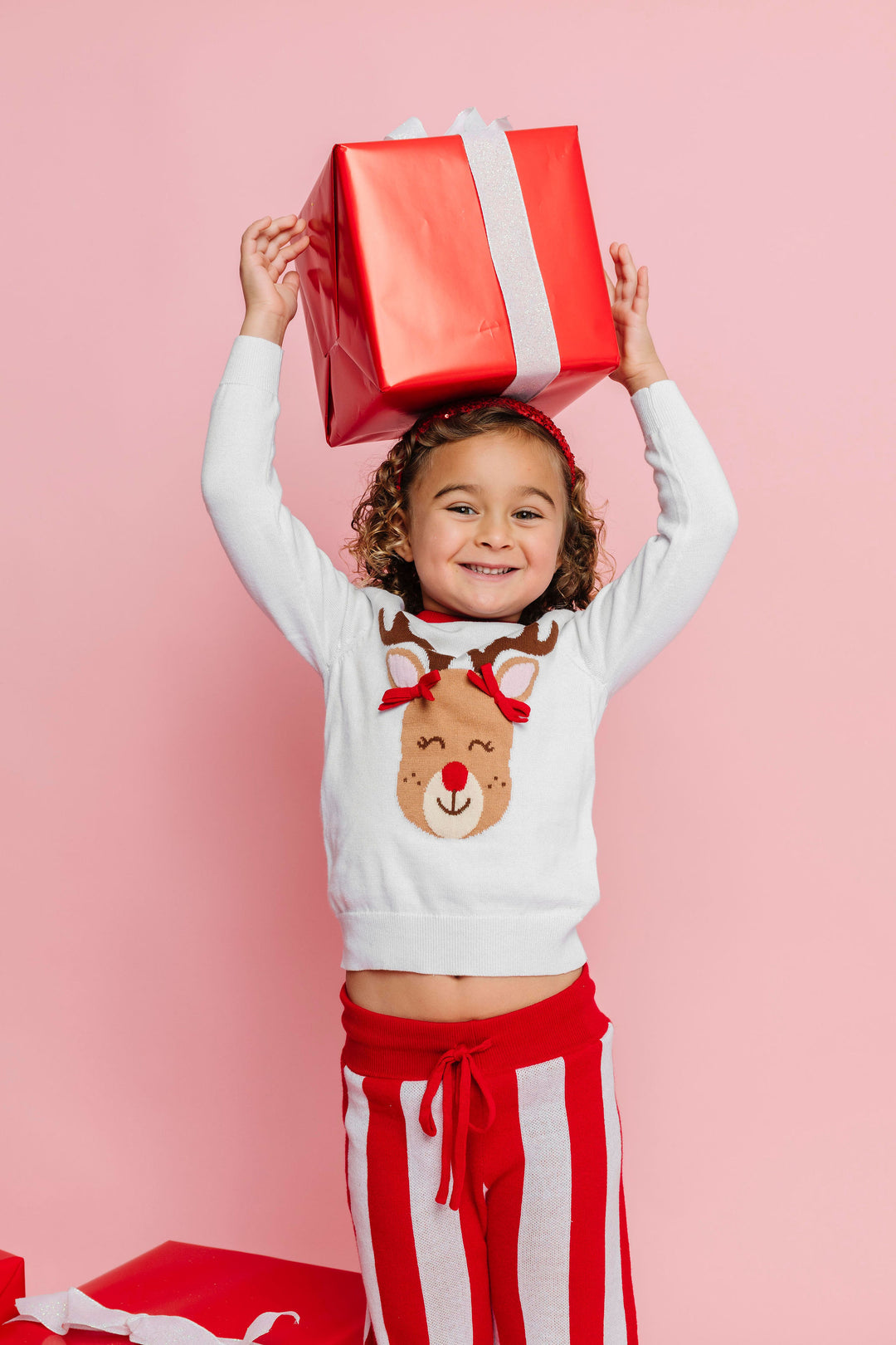 little girl holding Christmas gift in Rudolph sweater with red and pink striped sweater pants