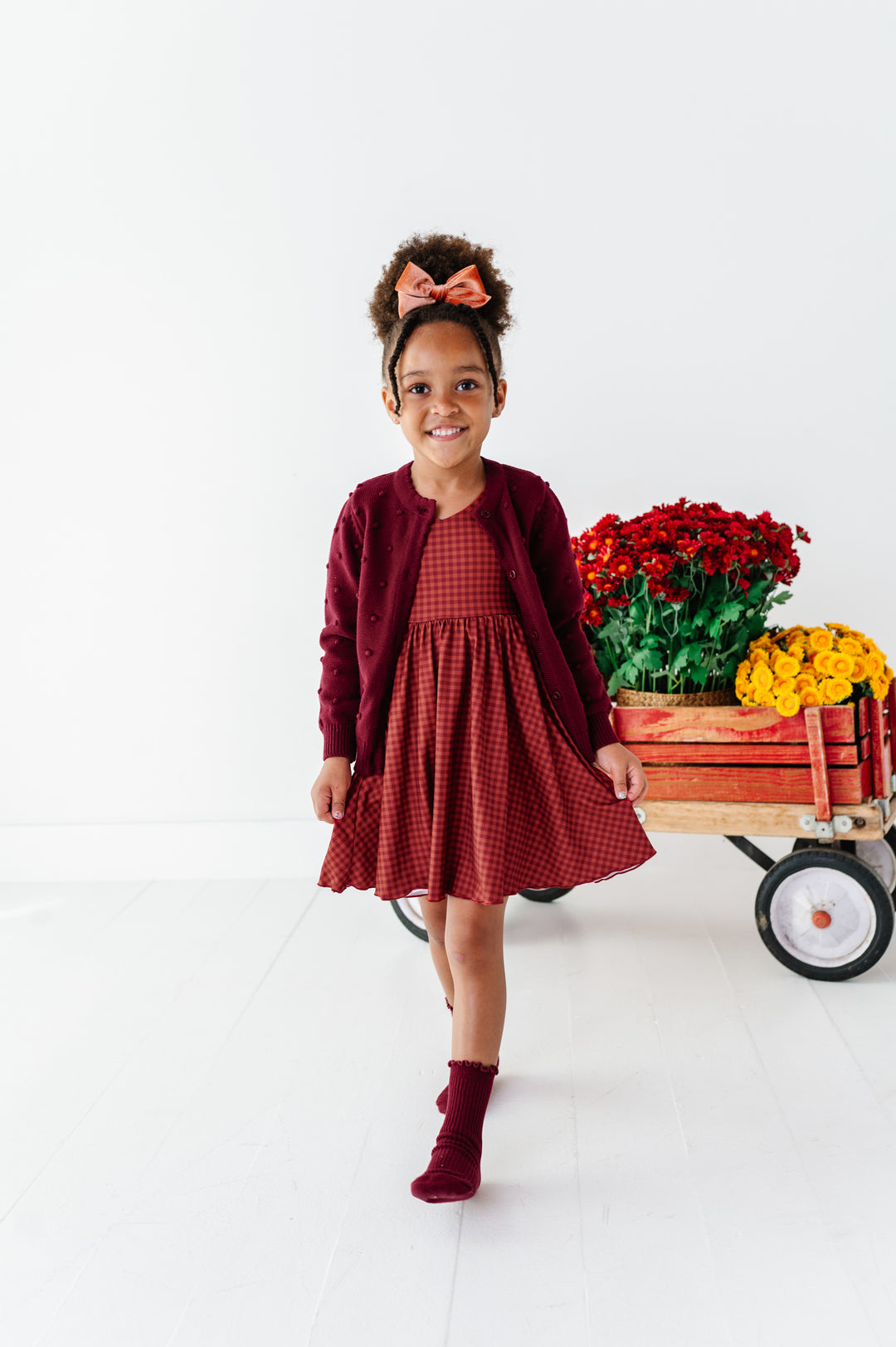 young girl with flower wagon in rust gingham fall twirl dress paired with burgundy cardigan and midi socks and coordinating velvet hair bow
