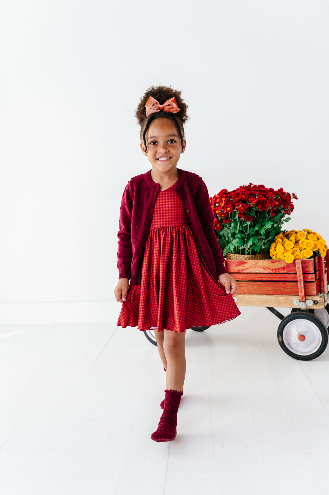 young girl with flower wagon in rust gingham fall twirl dress paired with burgundy cardigan and midi socks and coordinating velvet hair bow