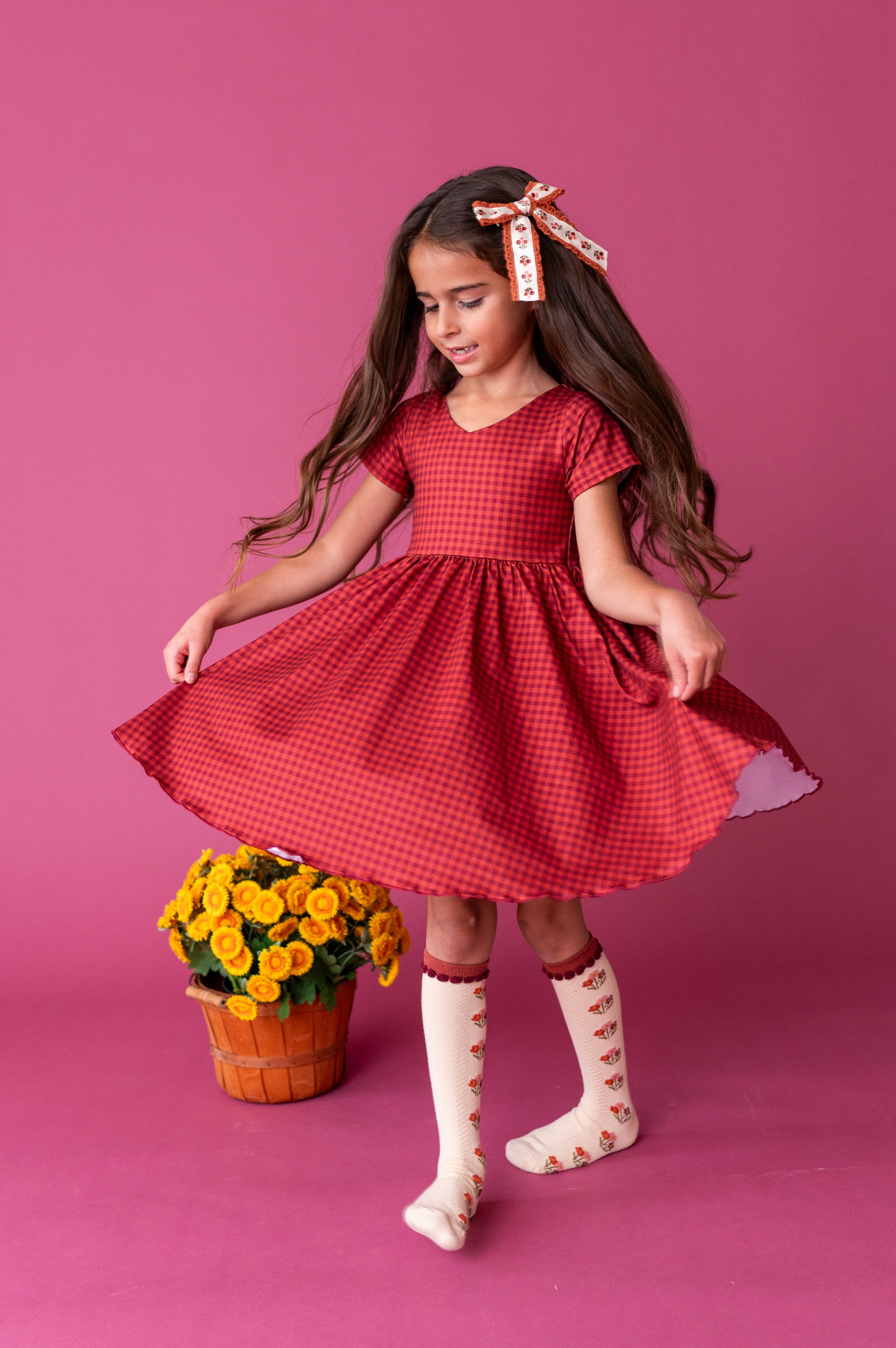 girl twirling by fall flowers in rust V-neck gingham print fall dress paired with cream knee socks with rust flowers and matching floral hair bow