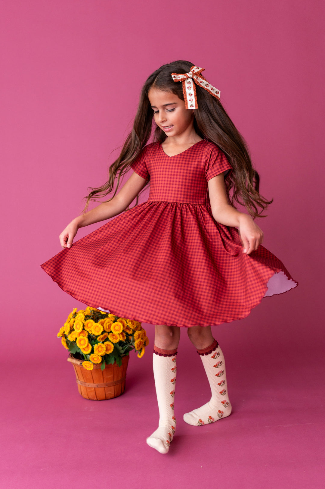 girl twirling by fall flowers in rust V-neck gingham print fall dress paired with cream knee socks with rust flowers and matching floral hair bow