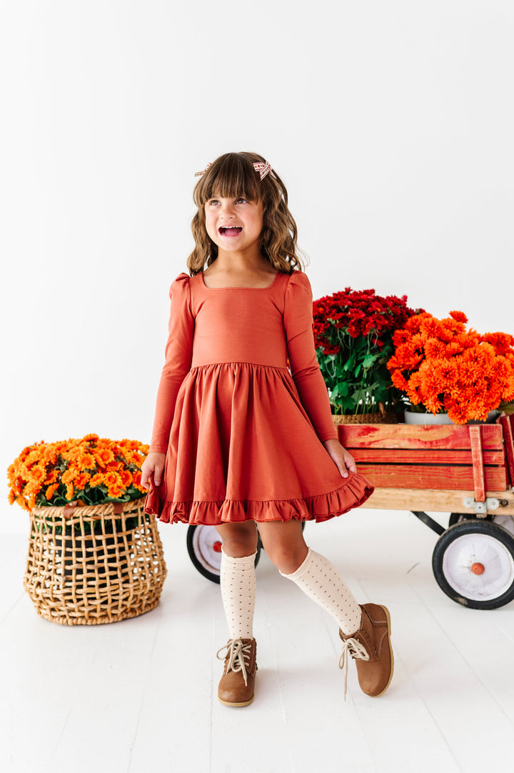 young girl in front of wagon filled with fall flowers in rust long sleeve twirl dress paired with cream knee socks with brown dot design
