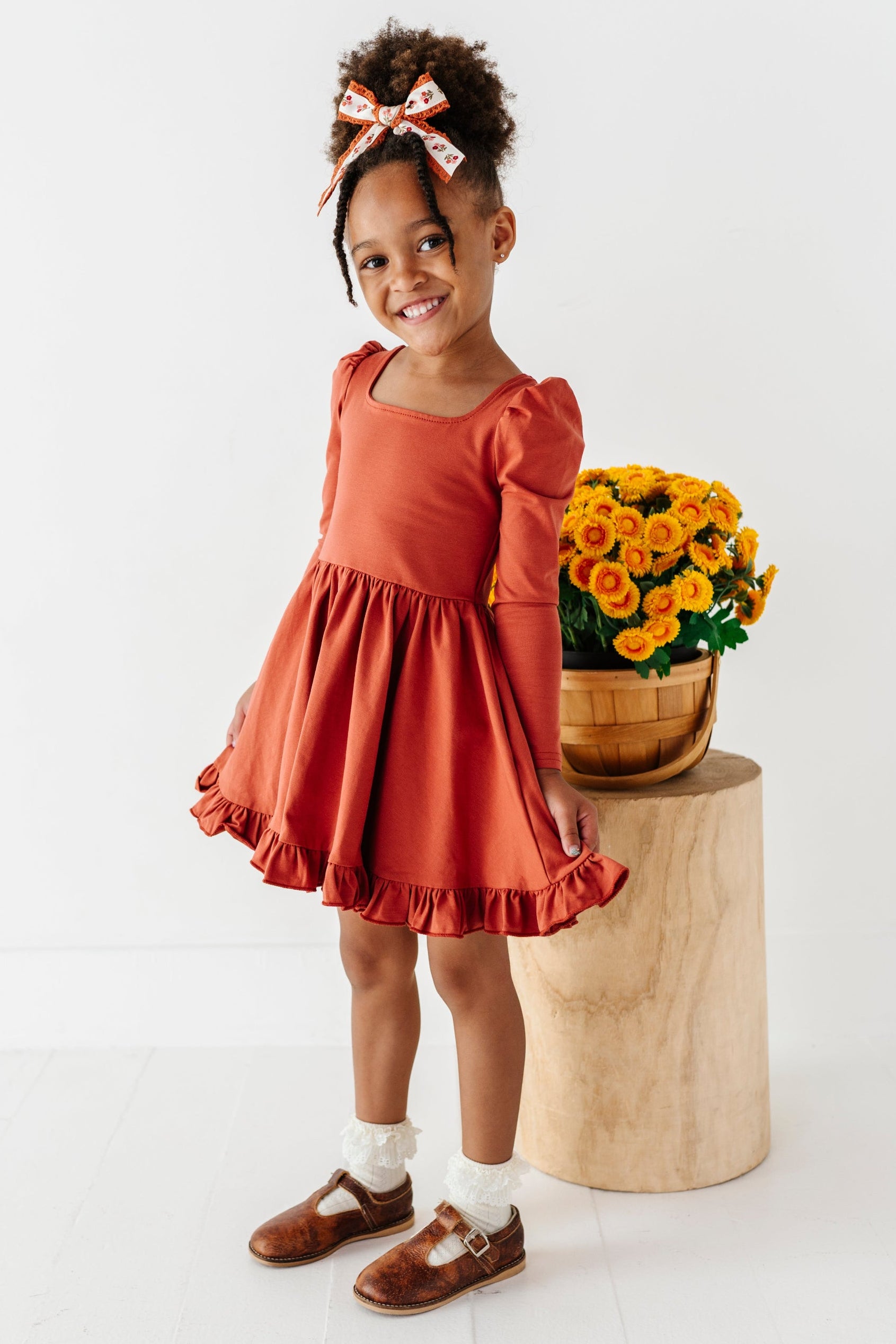 little girl in front of yellow fall flowers in rust long sleeve twirl dress paired with ivory lace bobby socks and rust floral hair bow