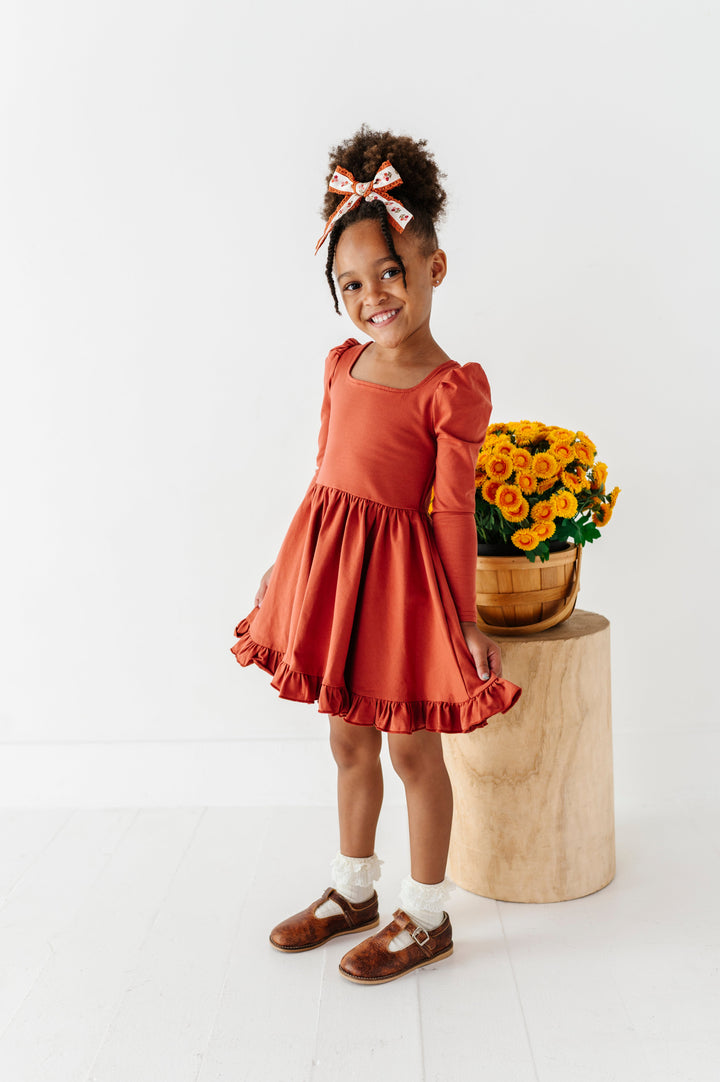 little girl in front of yellow fall flowers in rust long sleeve twirl dress paired with ivory lace bobby socks and rust floral hair bow